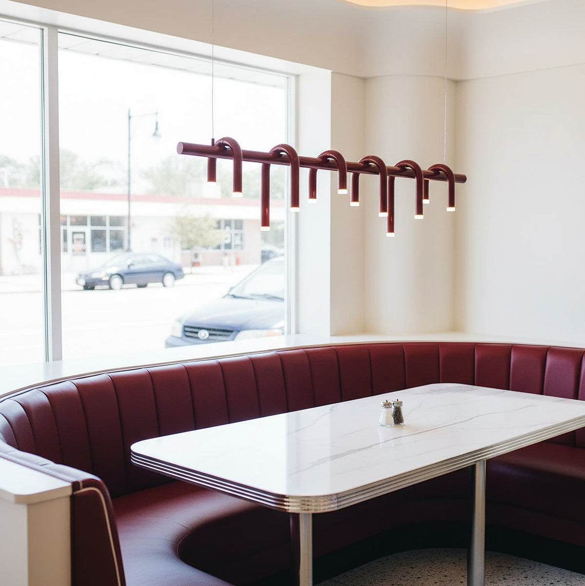 A linear red Clérachic chandelier hangs near a window above a white table and burgundy curved booth seating.