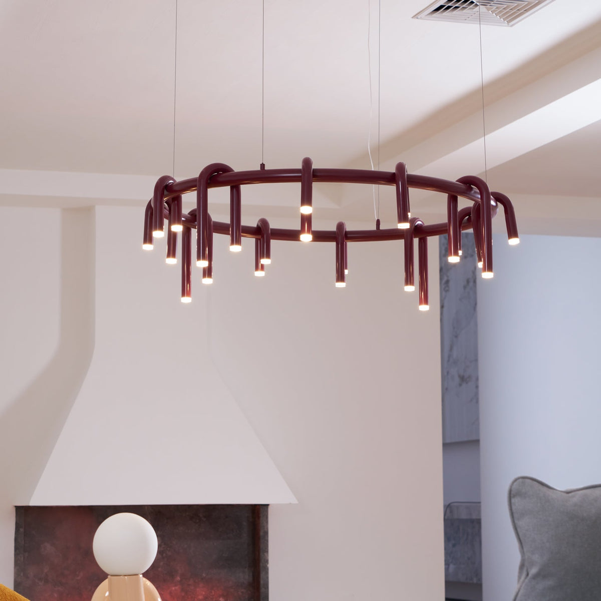 Red Clérachic ring chandelier in a white room, hung in front of a plaster fireplace hood with a small round-headed lamp below.