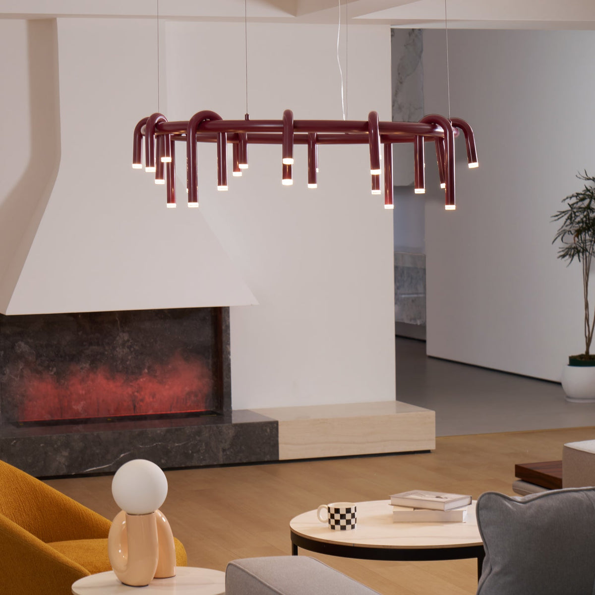 Red Clérachic ring chandelier seen at a slight angle in front of a white fireplace, with a plant and wooden floor beyond.