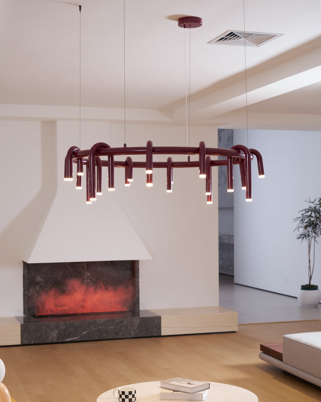 A red Clérachic ring chandelier hangs in an open room with a lit fireplace and a ceiling vent, shown from a low angle.