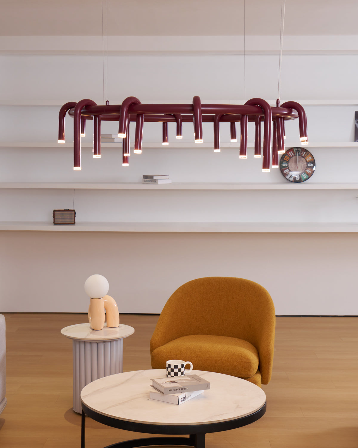 Ring chandelier in red from Clérachic, with a dense set of downward heads, hung in front of white wall shelves and an ochre chair.