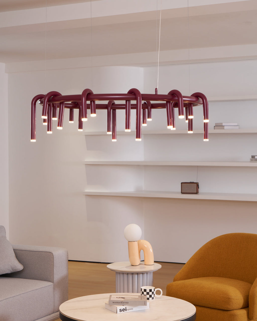 Red Clérachic ring chandelier above a small table with a sculptural ornament, beside a mustard armchair and a grey sofa.