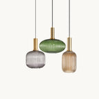 Three Clérachic reeded glass pendants in smoke grey, green and amber hang at different heights from gold fittings on a plain white backdrop.