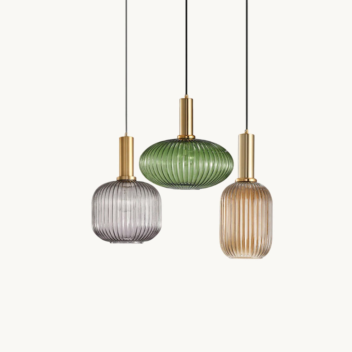 Three Clérachic reeded glass pendants in smoke grey, green and amber hang at different heights from gold fittings on a plain white backdrop.