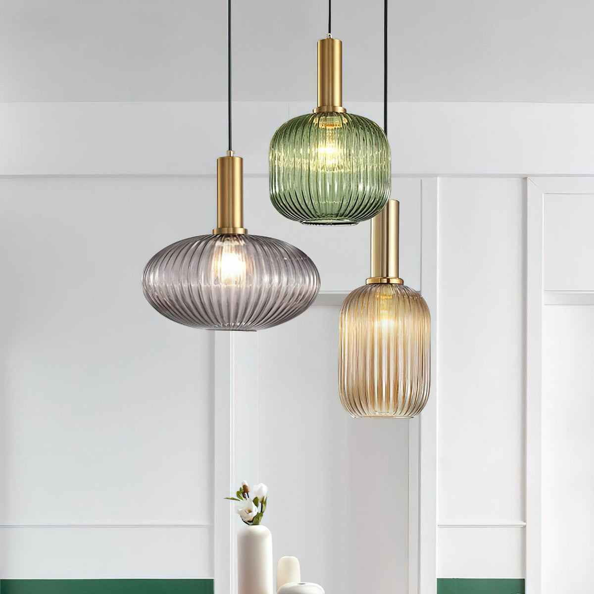 A trio of Clérachic pendants in smoke grey, green and amber glass, lit, hung in front of white panelled walls beside a small vase.