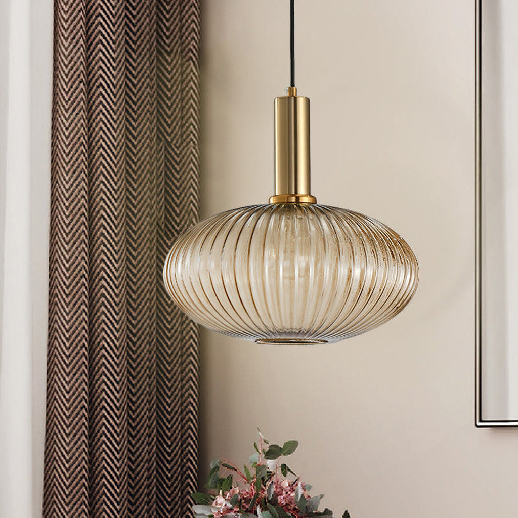 A wide, flattened amber reeded glass shade from Clérachic on a gold fitting, hanging beside a patterned curtain in a warm-toned room.