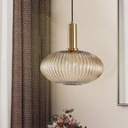A wide, flattened amber reeded glass shade from Clérachic on a gold fitting, hanging beside a patterned curtain in a warm-toned room.