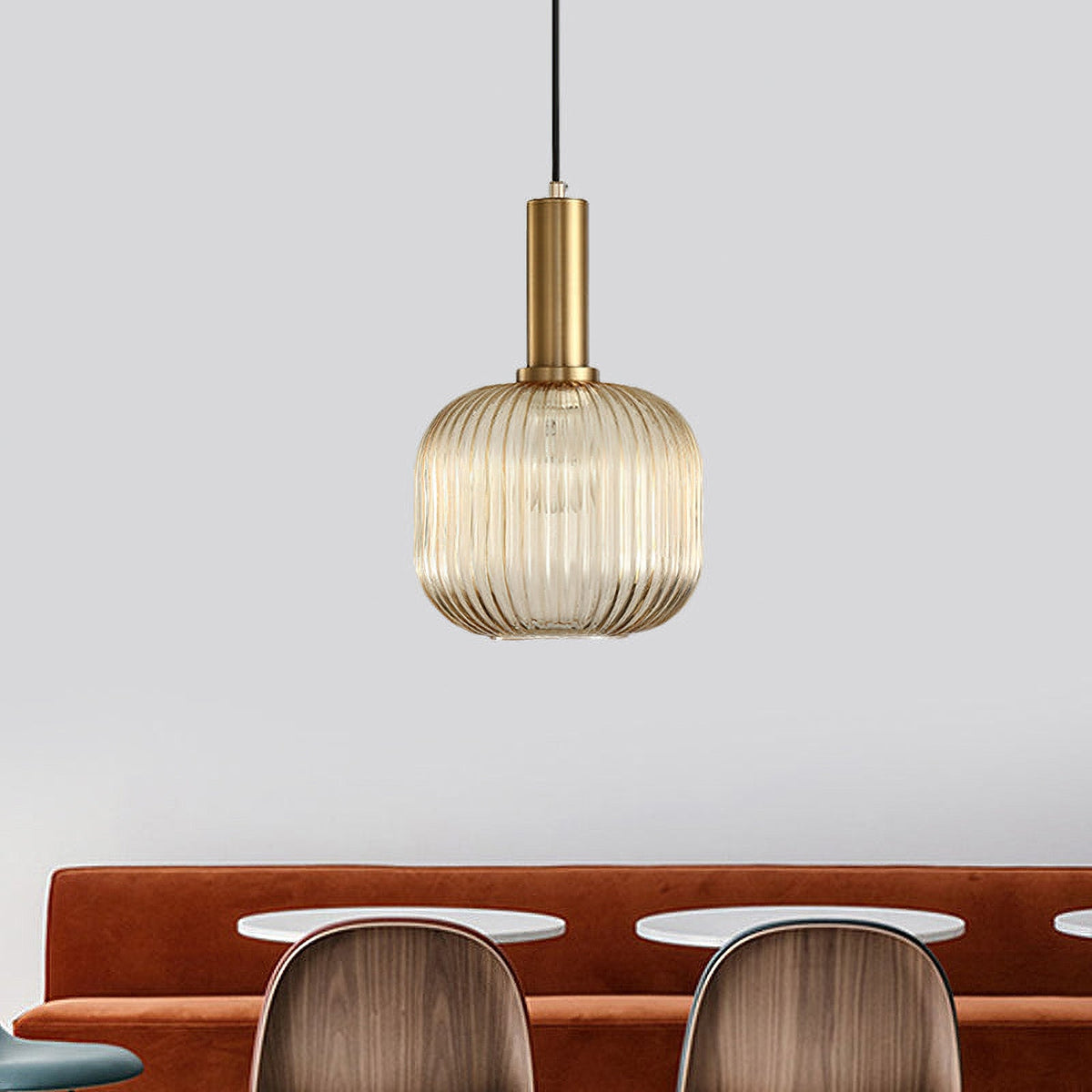This Clérachic pendant has a small rounded amber glass shade and gold fitting, hung against a pale grey wall above a rust-coloured banquette.