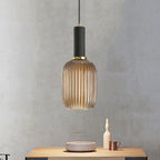 Tall, narrow amber reeded glass shade with a black fitting and gold ring, lit, from Clérachic, hanging above a light wooden table with ceramics.
