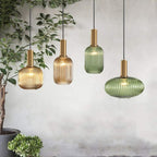 Four Clérachic reeded glass pendants in amber and green, in tall and flat shapes, hang at staggered heights beside a leafy indoor plant.