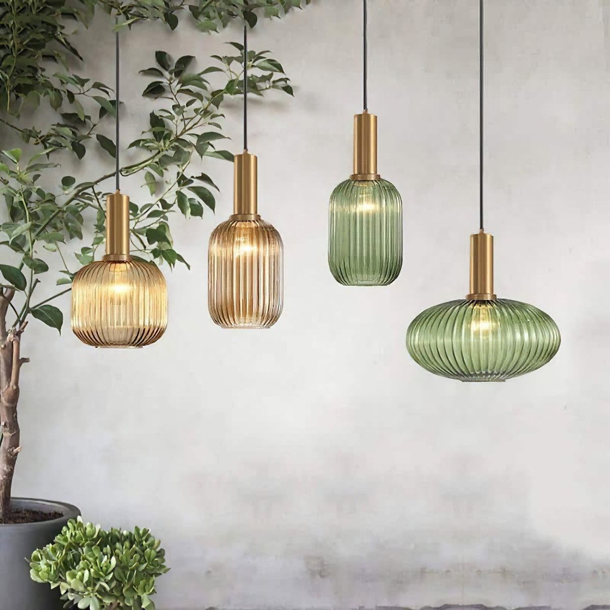 Four Clérachic reeded glass pendants in amber and green, in tall and flat shapes, hang at staggered heights beside a leafy indoor plant.