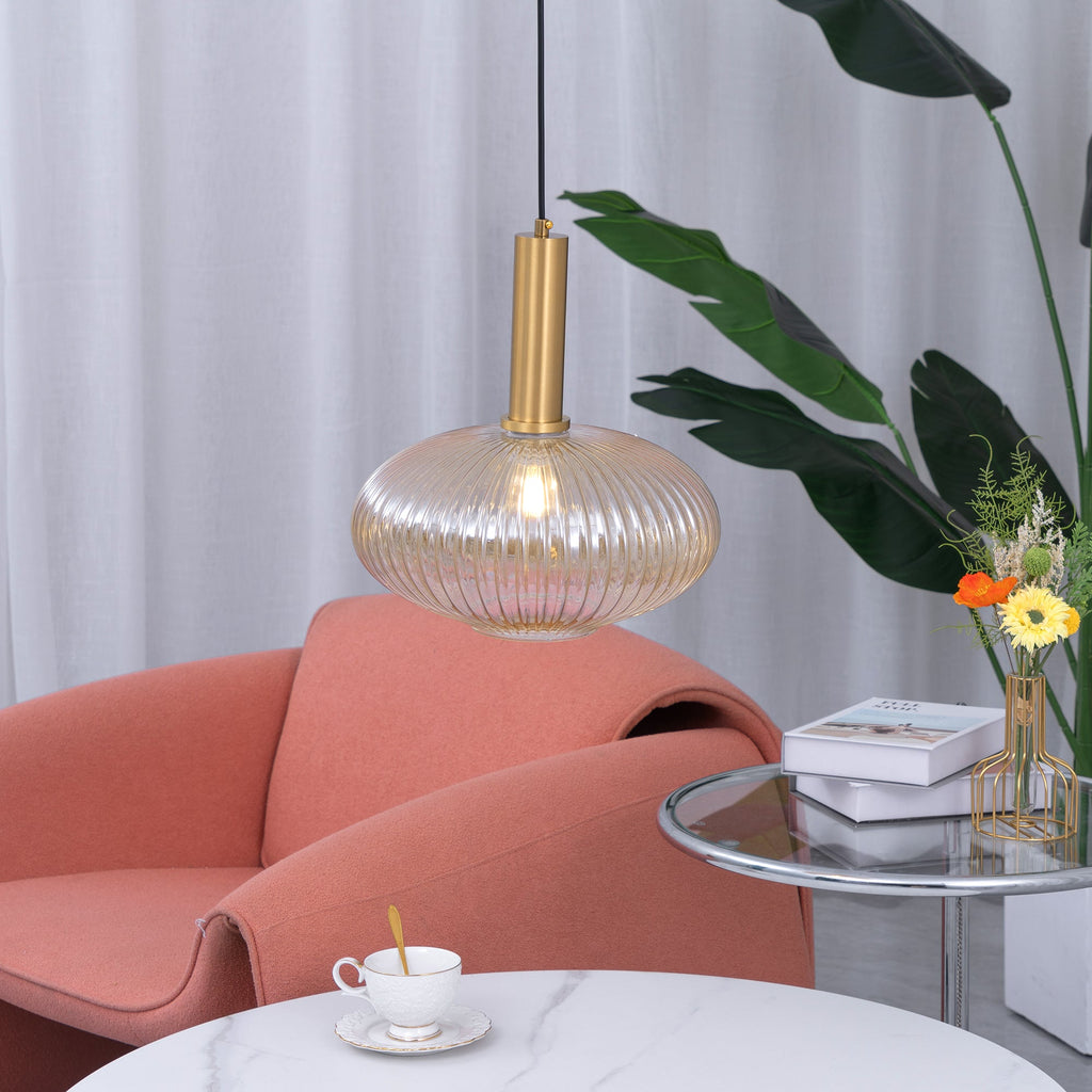 Lit amber glass pendant from Clérachic, wide and flat with fine vertical reeding, hanging over a white marble table beside a coral armchair.