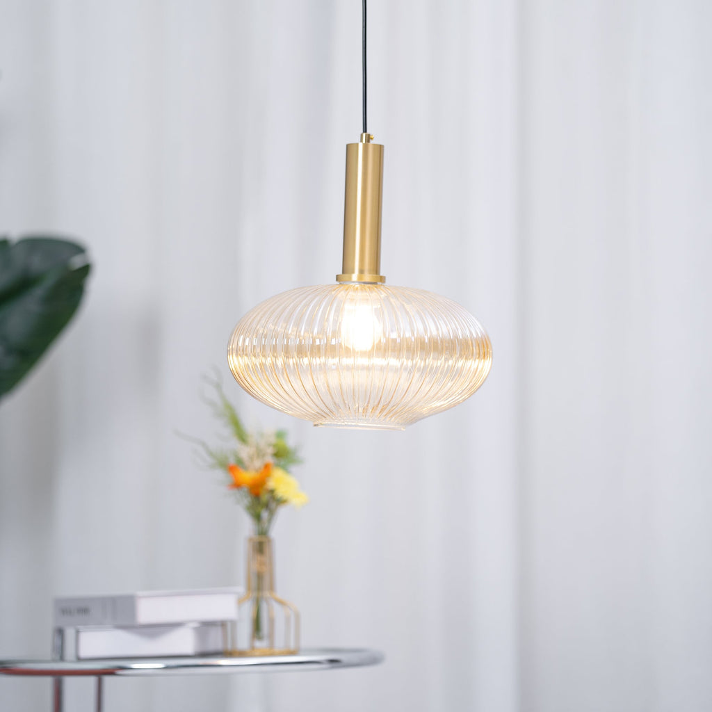 Close view of the lit amber reeded glass shade and gold stem of this Clérachic pendant, with a vase of orange flowers softly blurred behind.