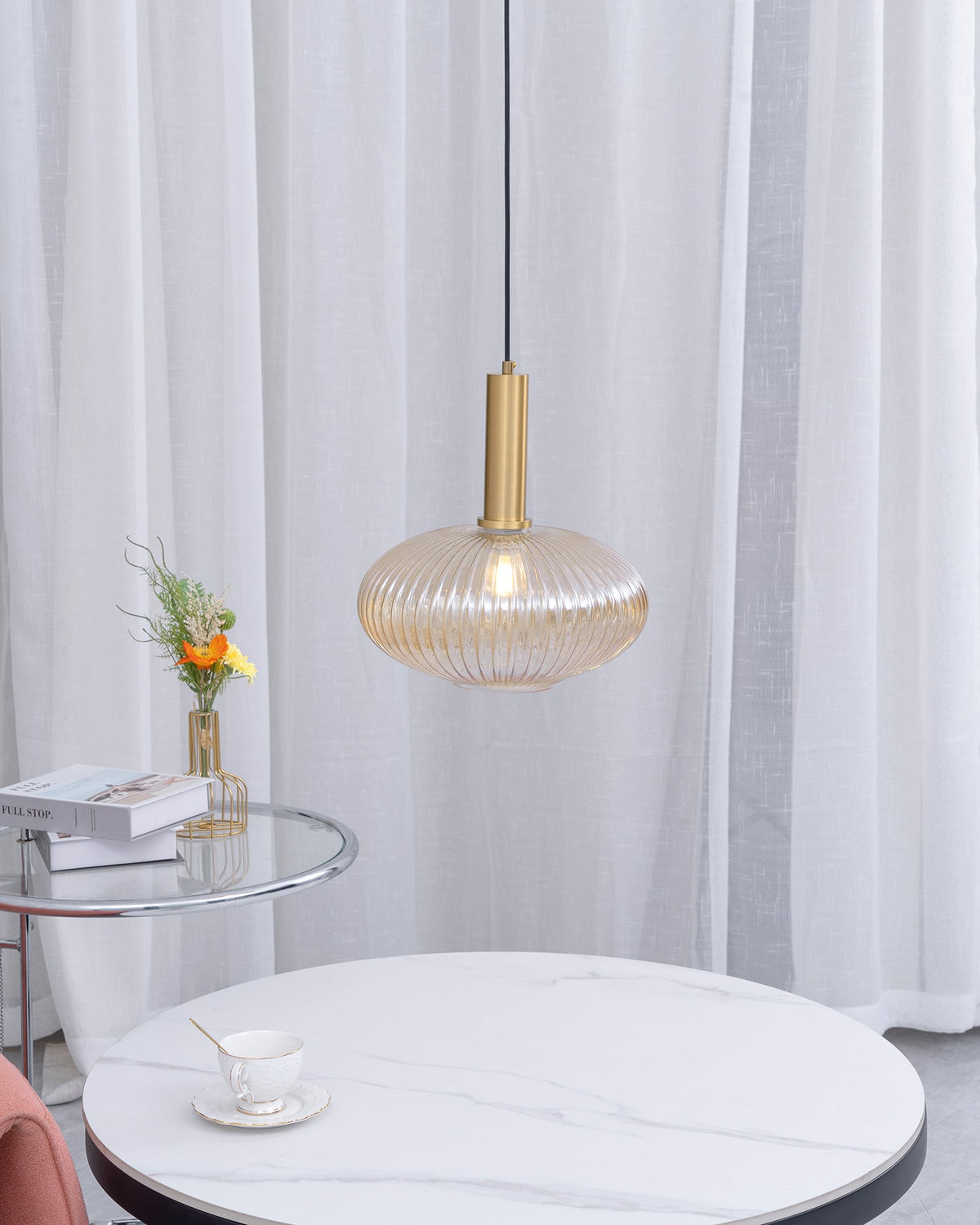 A small flat amber reeded pendant from Clérachic hangs in front of sheer white curtains above a round white marble coffee table.