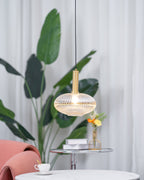 Gold-stemmed Clérachic pendant with a lit, flattened amber reeded shade, set in front of a banana-leaf plant and sheer curtains.