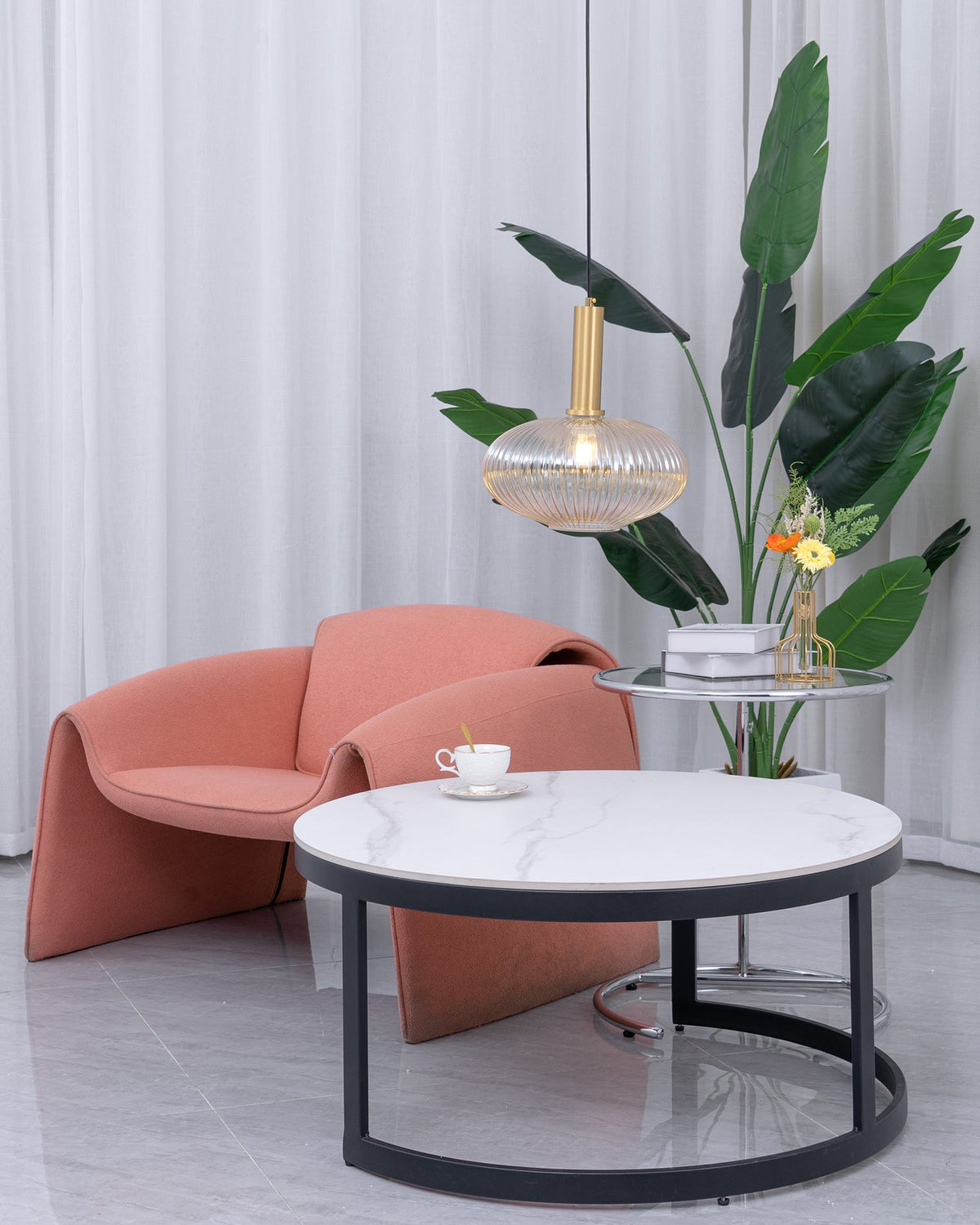 Wide view of a living corner where a small amber reeded Clérachic pendant hangs near a banana-leaf plant, a coral armchair and a marble table.