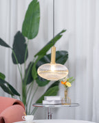 Lit amber reeded glass pendant from Clérachic hanging in front of tall green leaves, with a teacup and flowers on a table below.