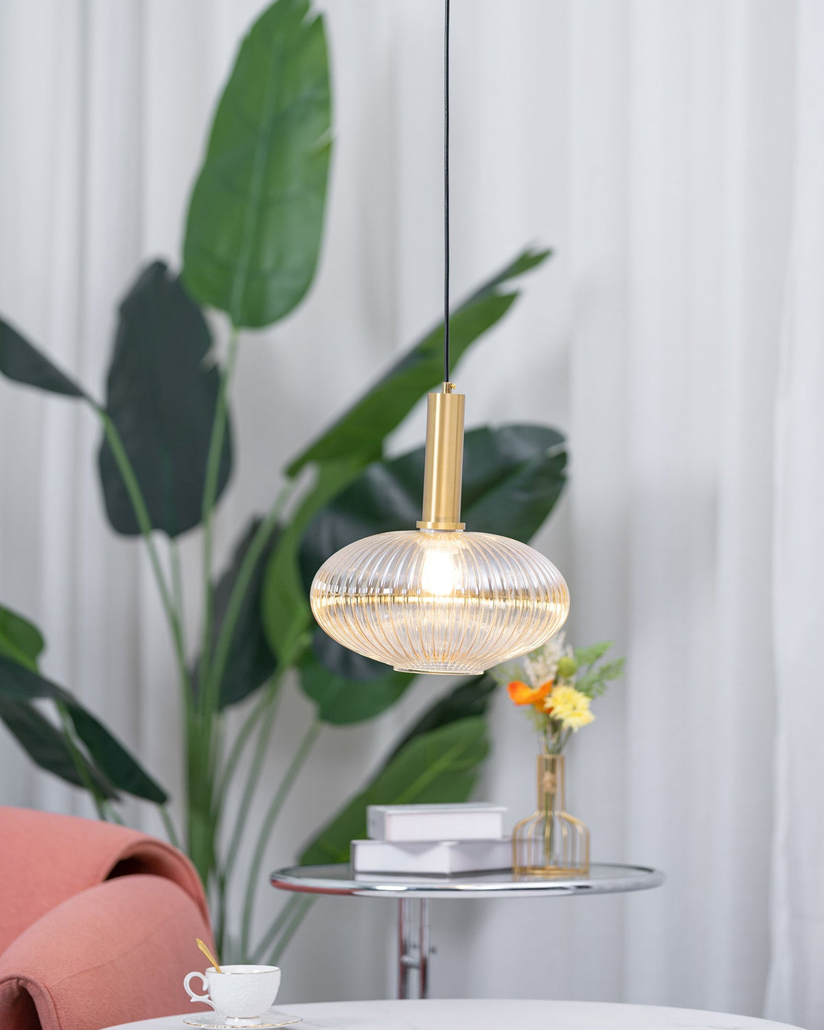 Lit amber reeded glass pendant from Clérachic hanging in front of tall green leaves, with a teacup and flowers on a table below.