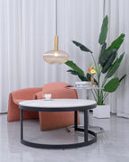 Small amber Clérachic pendant with a gold stem hangs above a round marble coffee table, with a pink armchair and a large plant beyond.