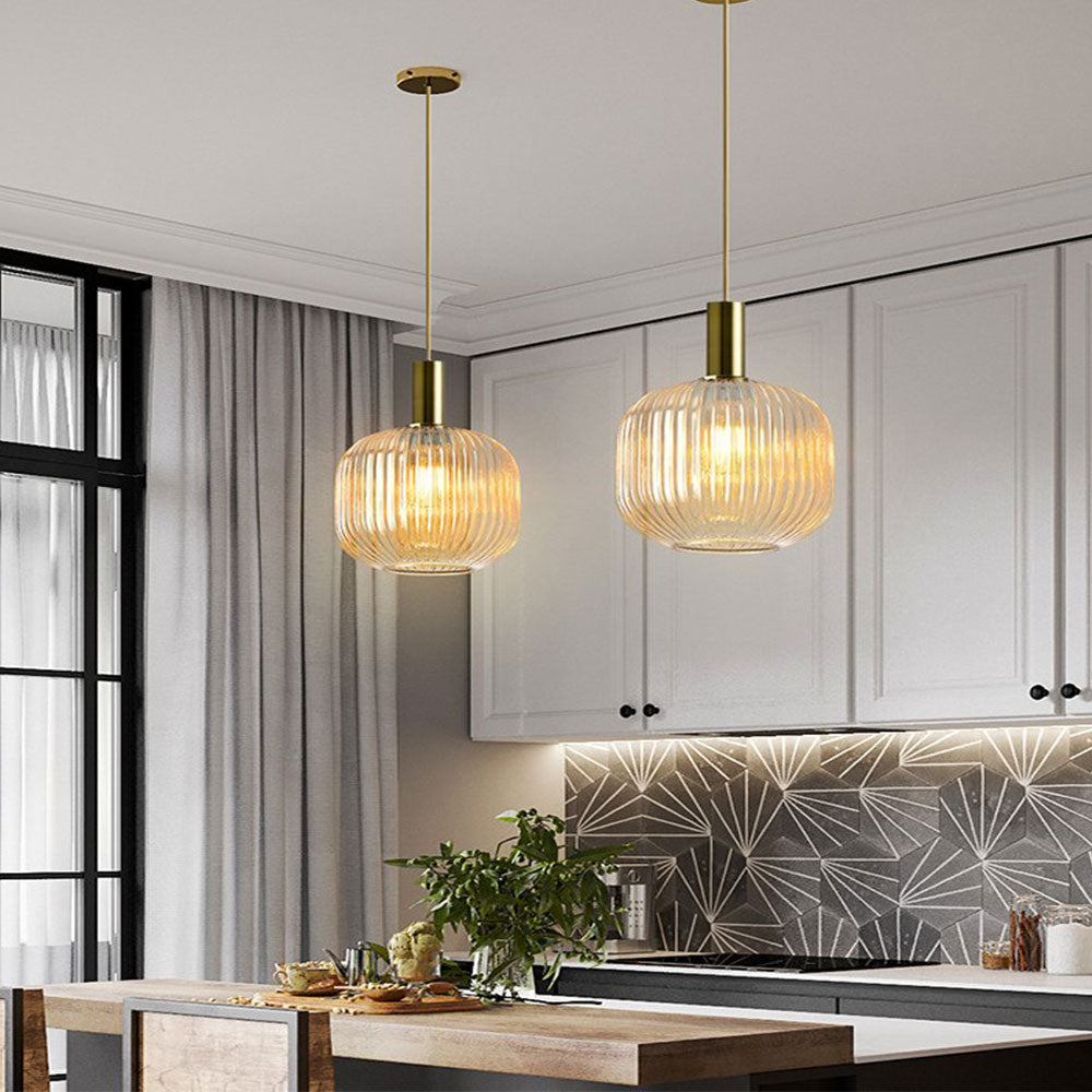 Two Clérachic pendants with tall amber reeded glass shades hang over a kitchen island in front of white cabinets and a patterned tile splashback.