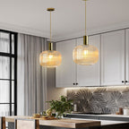 Two Clérachic pendants with tall amber reeded glass shades hang over a kitchen island in front of white cabinets and a patterned tile splashback.