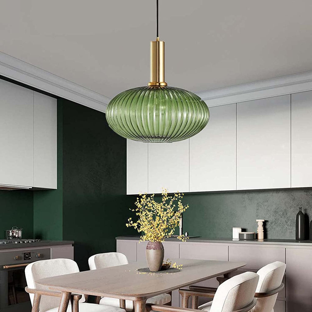 A flat green reeded glass pendant from Clérachic on a gold stem hangs in a kitchen with dark green walls, white cabinets and a wooden table.