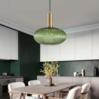 A flat green reeded glass pendant from Clérachic on a gold stem hangs in a kitchen with dark green walls, white cabinets and a wooden table.