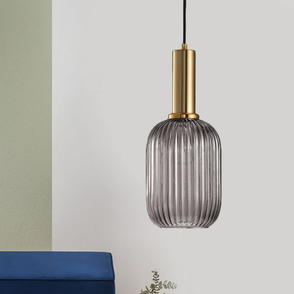 Tall smoke grey reeded glass shade with a gold stem from Clérachic, hanging against a pale wall with a blue cushion at the bottom edge.