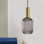 Tall smoke grey reeded glass shade with a gold stem from Clérachic, hanging against a pale wall with a blue cushion at the bottom edge.