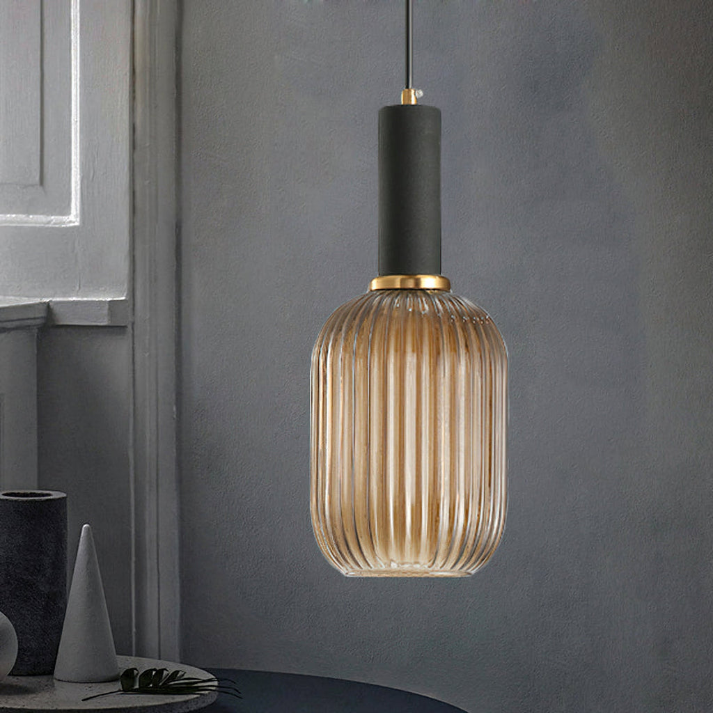 Tall amber reeded glass shade from Clérachic with a black fitting, hanging in front of a dark grey wall with a grey vase and cone nearby.