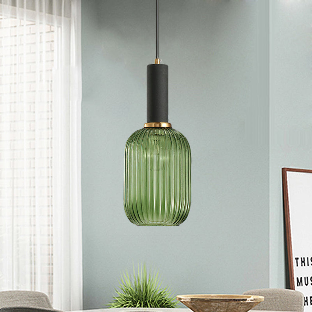 Tall green reeded glass pendant from Clérachic with a black fitting hangs against a soft grey-green wall, with a wooden bowl and plant below.