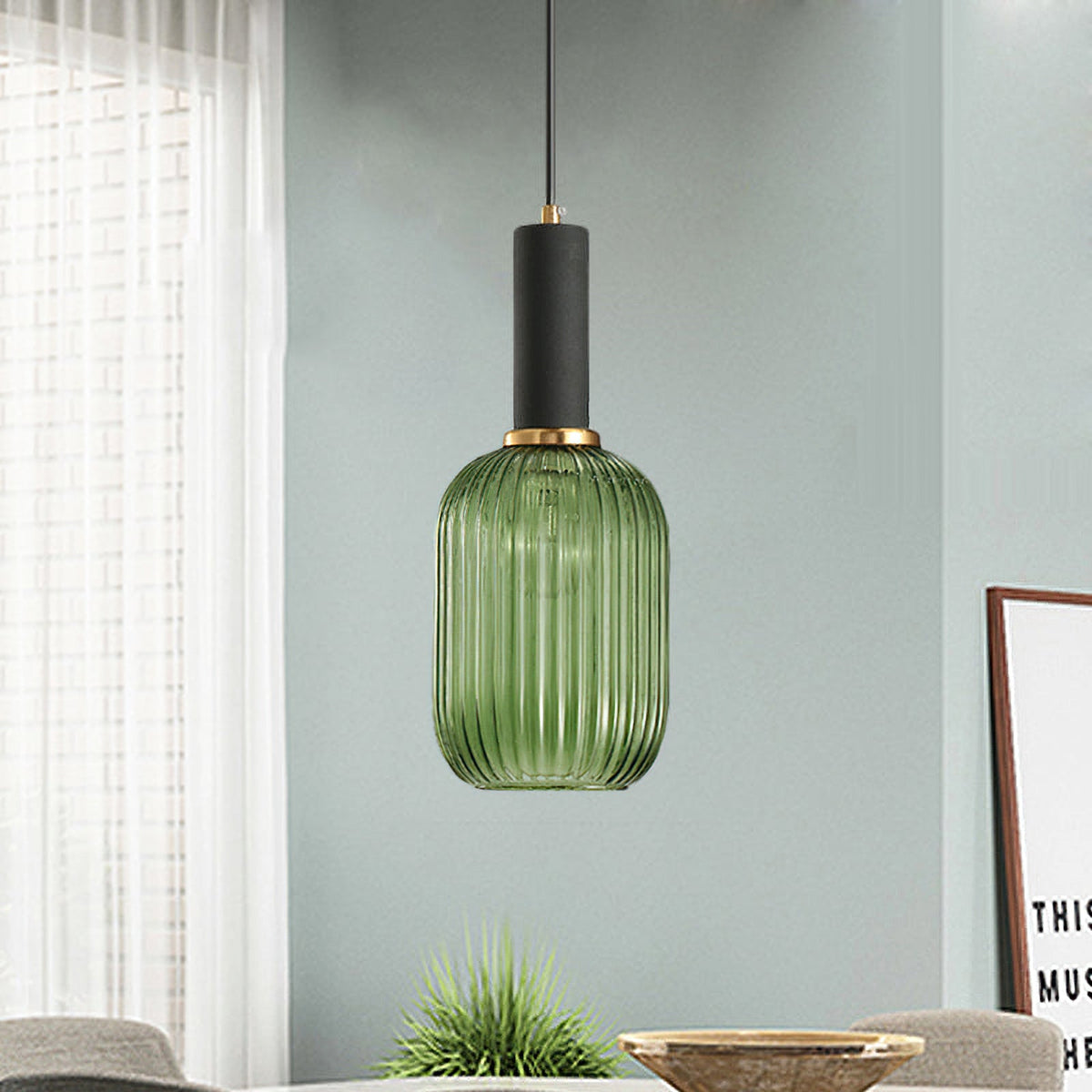 Tall green reeded glass pendant from Clérachic with a black fitting hangs against a soft grey-green wall, with a wooden bowl and plant below.