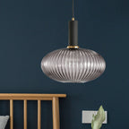A wide smoke grey reeded glass pendant from Clérachic with a black fitting hangs against a deep navy wall beside a wooden chair back.