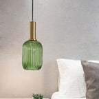 Tall green reeded glass pendant from Clérachic on a gold stem, hanging in front of a grey wall beside a white bed.