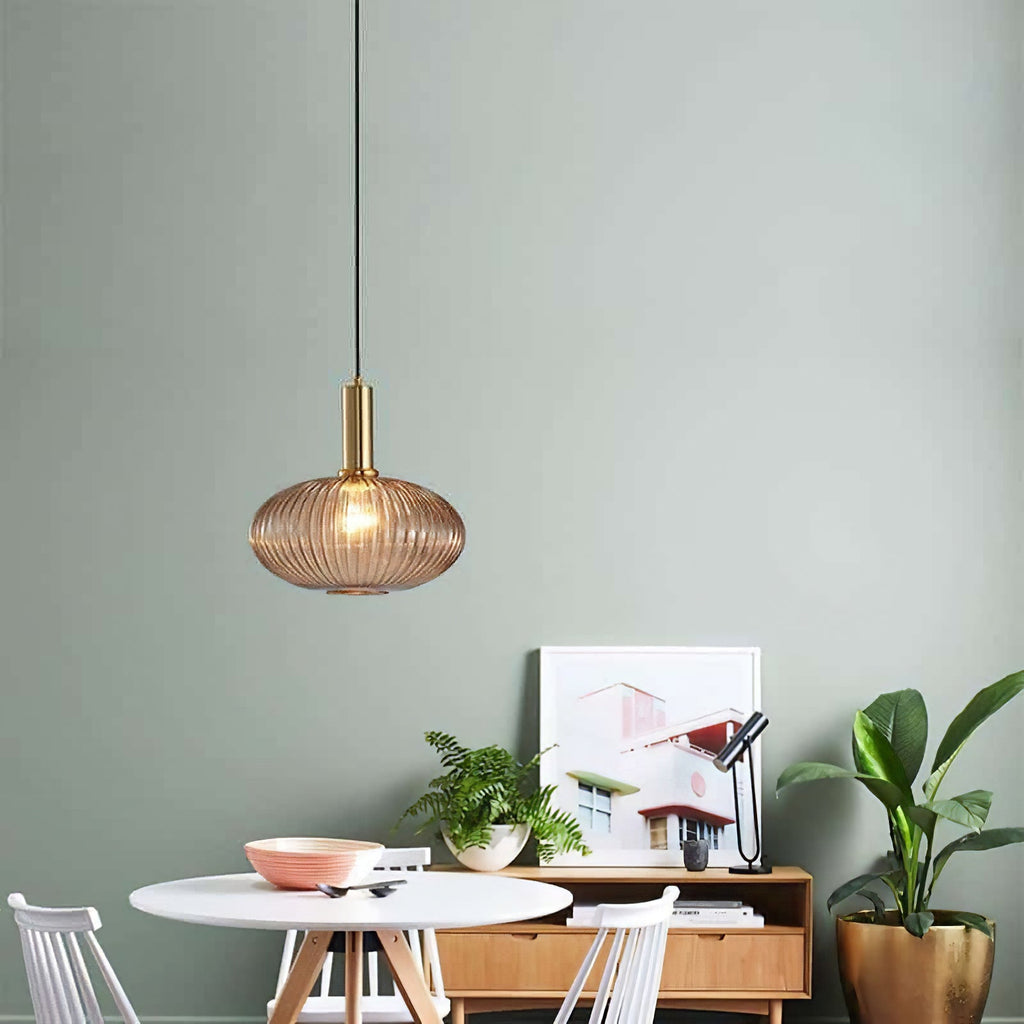 Small flat amber reeded glass pendant from Clérachic on a gold stem hangs against a sage-green wall above a round white table and sideboard.