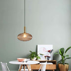 Small flat amber reeded glass pendant from Clérachic on a gold stem hangs against a sage-green wall above a round white table and sideboard.