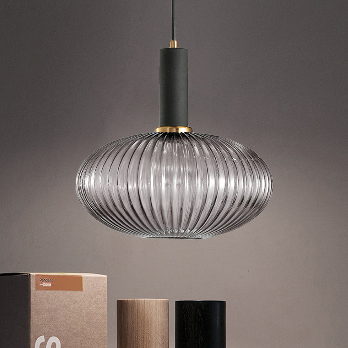 Wide smoke grey reeded glass pendant from Clérachic with a black fitting, against a taupe wall with a few boxes and wooden pots below.