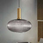 A flat smoke grey reeded glass pendant from Clérachic on a gold stem hangs against a grey wall with wooden slatted blinds beside it.