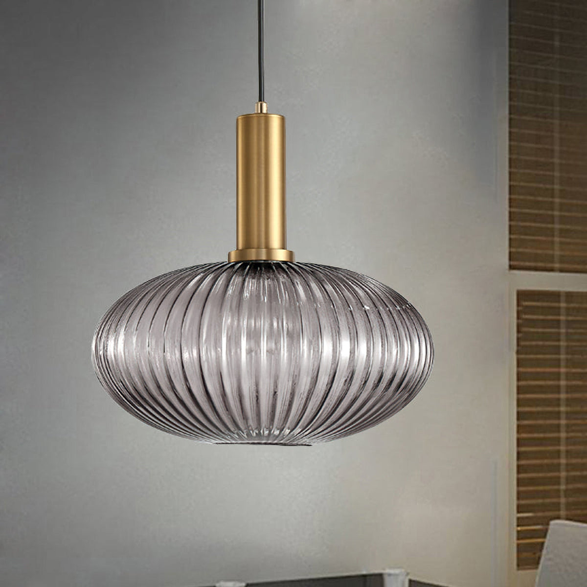 A flat smoke grey reeded glass pendant from Clérachic on a gold stem hangs against a grey wall with wooden slatted blinds beside it.