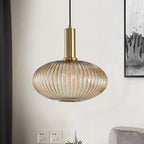 Wide amber reeded glass pendant from Clérachic on a gold stem hangs against a white wall with a framed print and wall sockets in view.