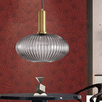 A wide smoke grey reeded glass pendant from Clérachic on a gold stem, hanging against a red patterned wall above a dark round table.