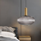 Smoke grey reeded glass pendant from Clérachic on a gold stem hangs beside a bed, with a wooden bedside table and a grey headboard.