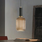 Tall amber reeded glass pendant from Clérachic with a black fitting hangs in a grey room with a dark wooden panel and books on a table.