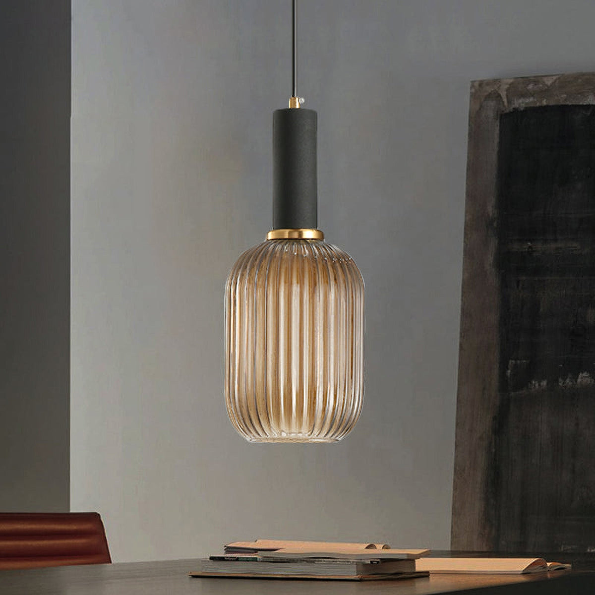 Tall amber reeded glass pendant from Clérachic with a black fitting hangs in a grey room with a dark wooden panel and books on a table.