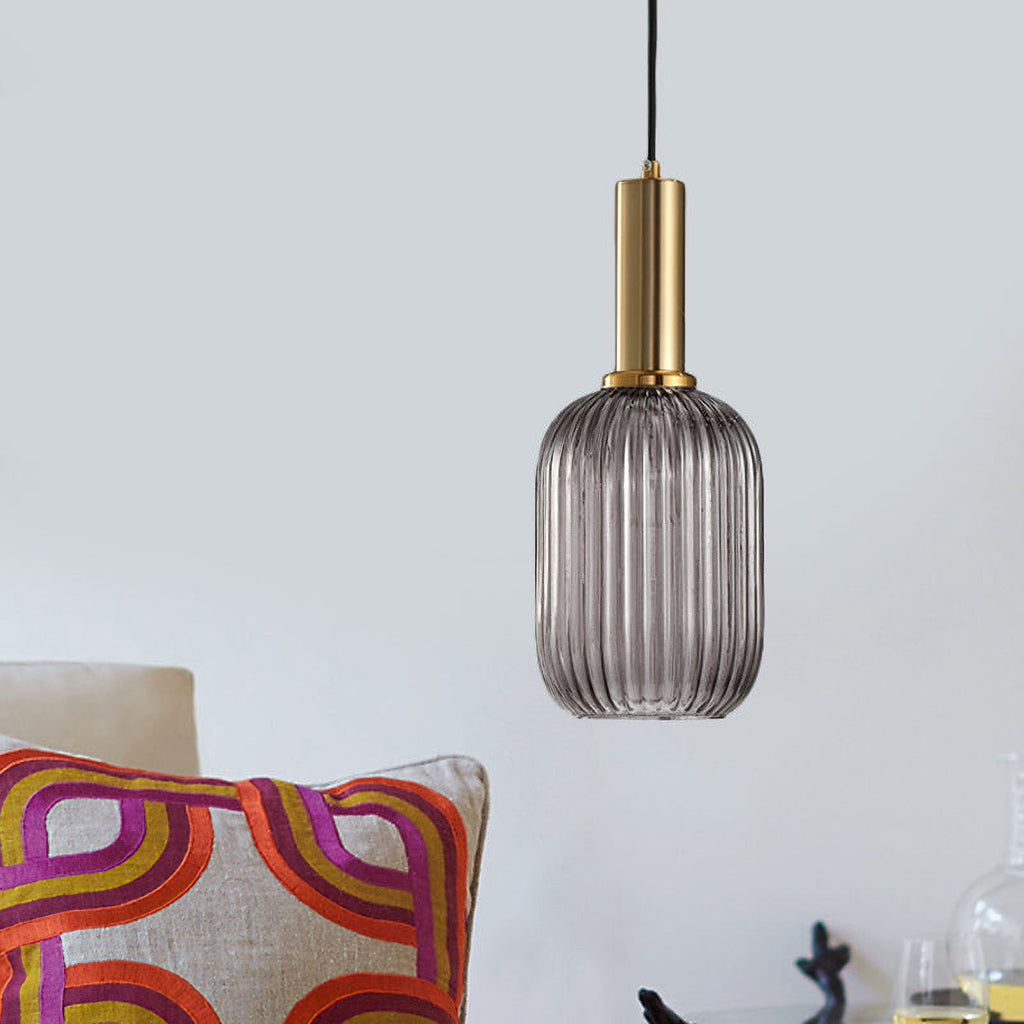 Smoke grey reeded glass pendant from Clérachic on a gold stem, hanging against a pale wall with a colourful patterned cushion below.