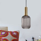 Smoke grey reeded glass pendant from Clérachic on a gold stem, hanging against a pale wall with a colourful patterned cushion below.