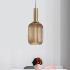 A tall amber reeded glass pendant from Clérachic on a gold stem hangs against a pale wall with a pot of dried flowers on a table.