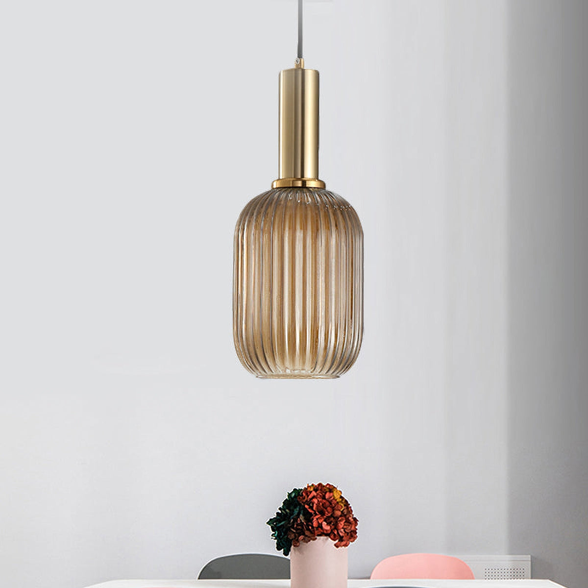 A tall amber reeded glass pendant from Clérachic on a gold stem hangs against a pale wall with a pot of dried flowers on a table.