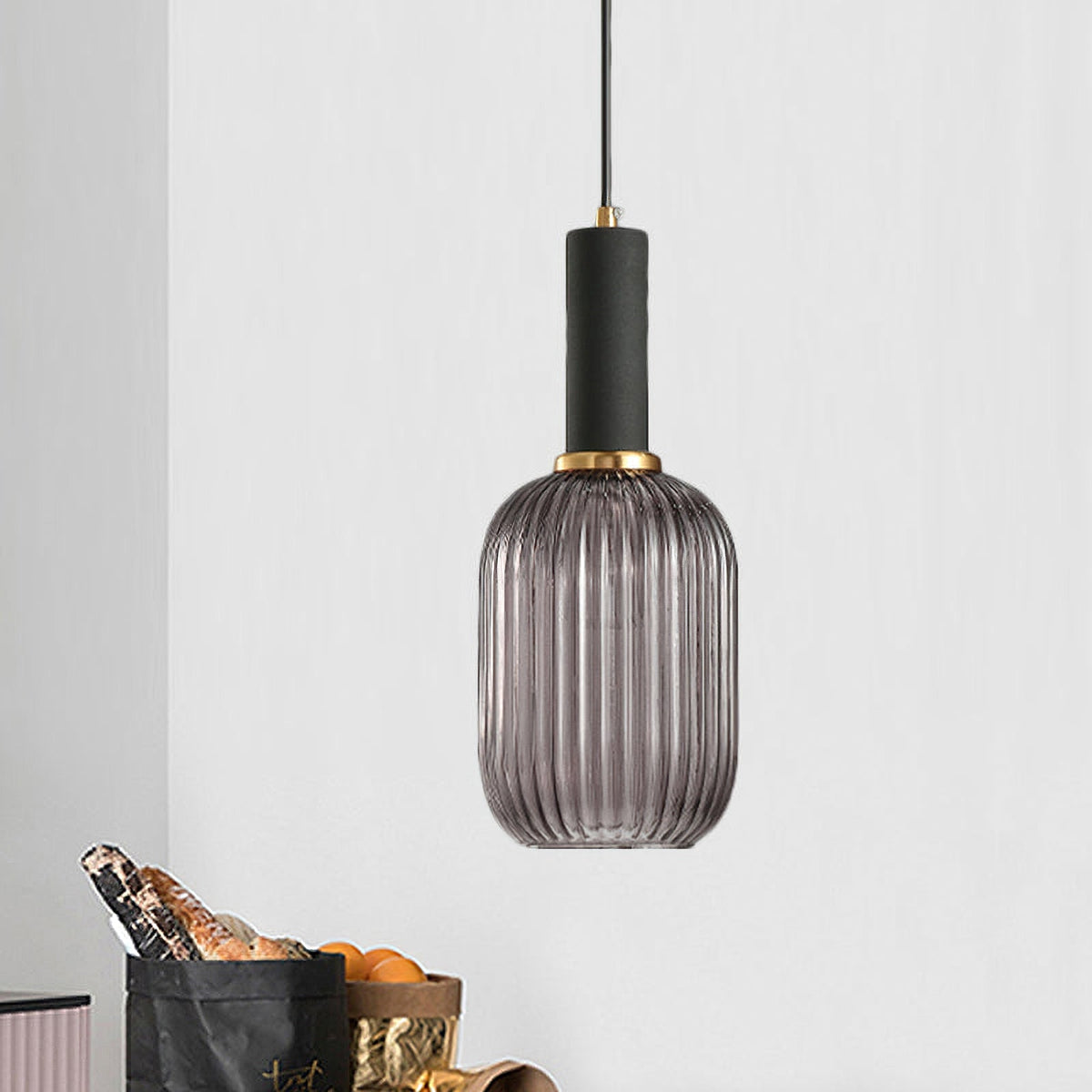 Tall smoke grey reeded glass pendant from Clérachic with a black fitting hangs in a pale room with a shopping bag and oranges below.