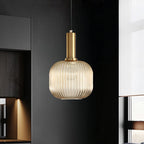 A round amber reeded glass pendant from Clérachic on a gold stem hangs against a dark kitchen unit, glowing softly.